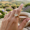 Woman's elegant hand wearing gold paved heart ring with champagne zircon stones, holding a floral tea cup with lush garden background