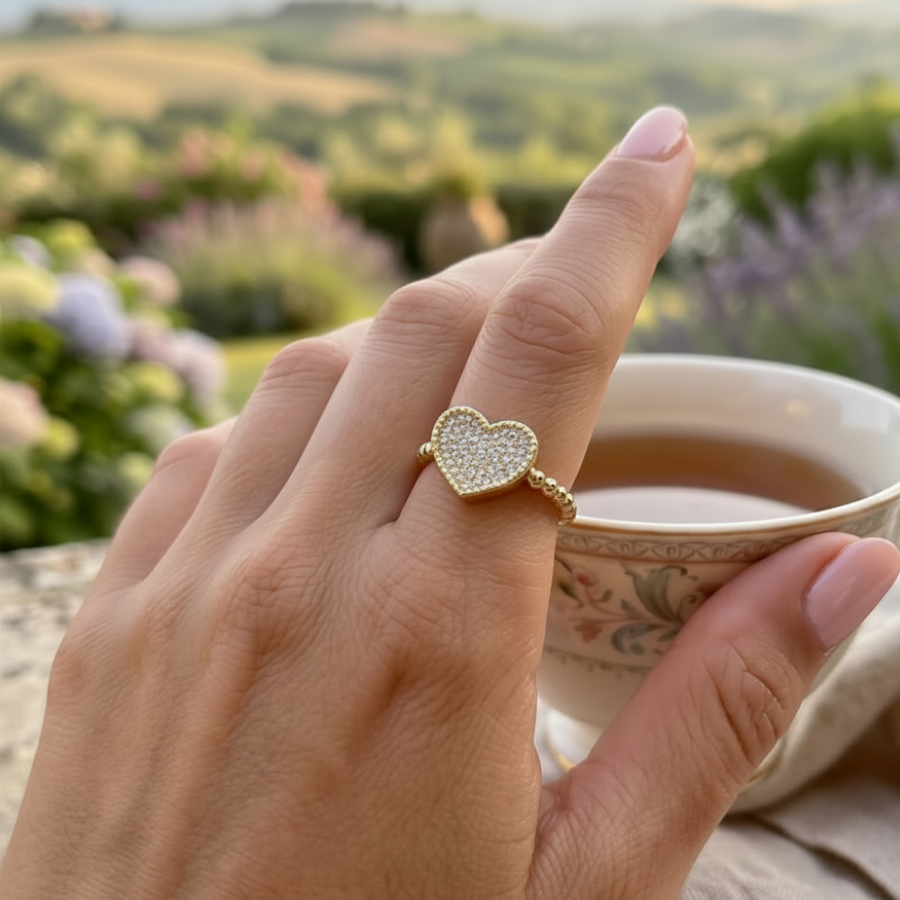 Woman's elegant hand wearing gold paved heart ring with champagne zircon stones, holding a floral tea cup with lush garden background